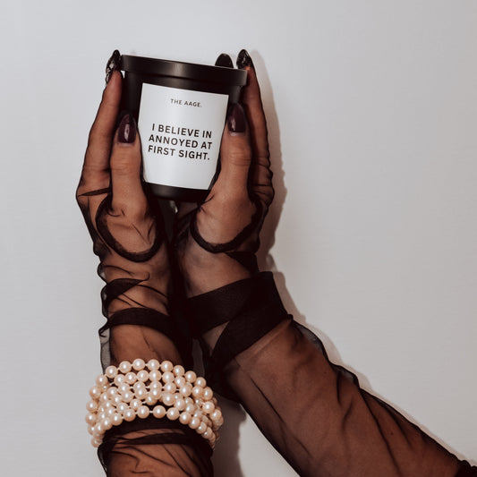 A person holding a candle in a black and white patterned holder, with a label on the candle that reads 'I BELIEVE IN ANNOYED AT FIRST SIGHT.'.